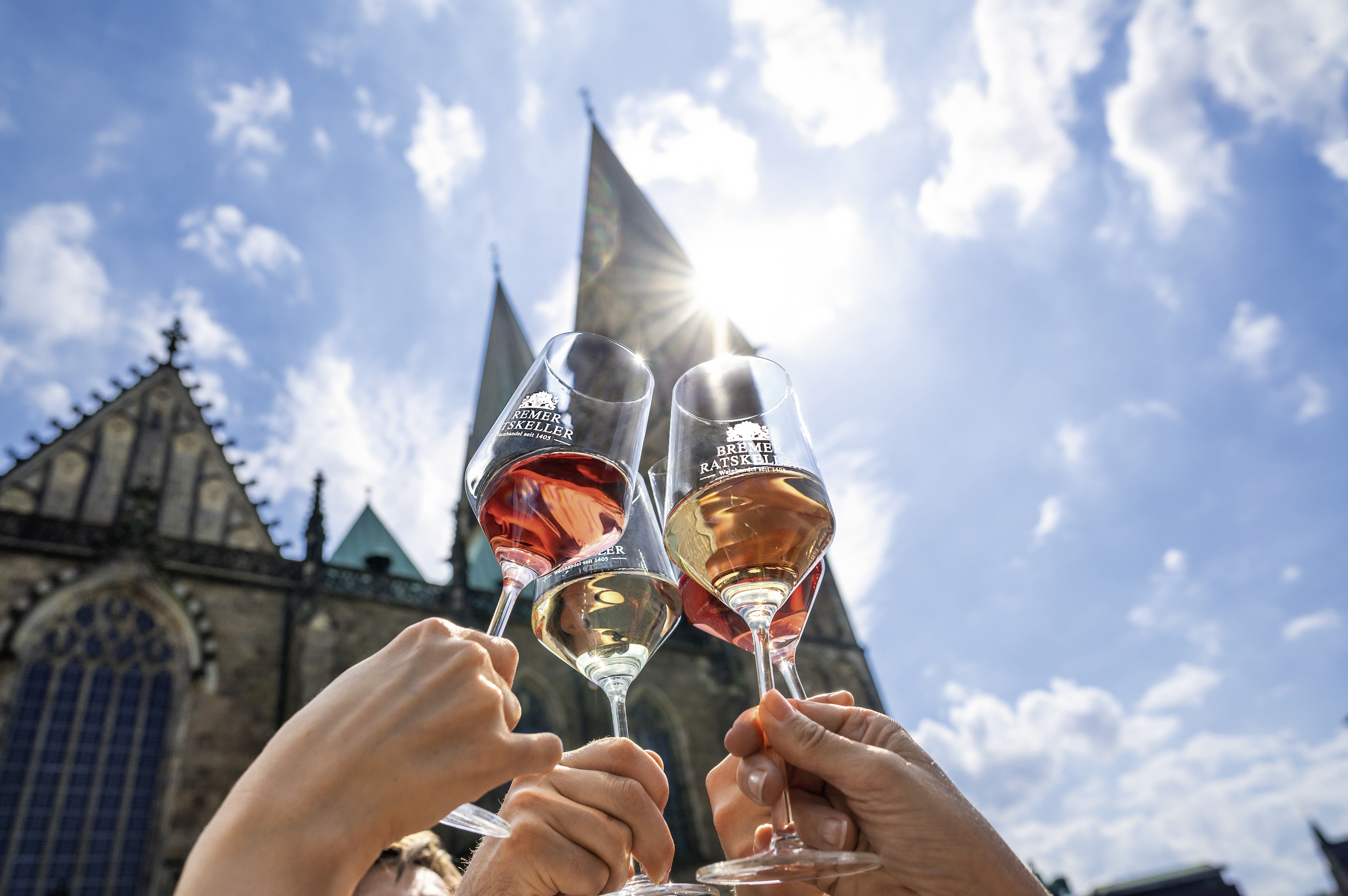Anstoßen mit Weingläsern vom Bremer Ratskeller vor blauem Himmel und dem Bremer Dom im Hintergrund