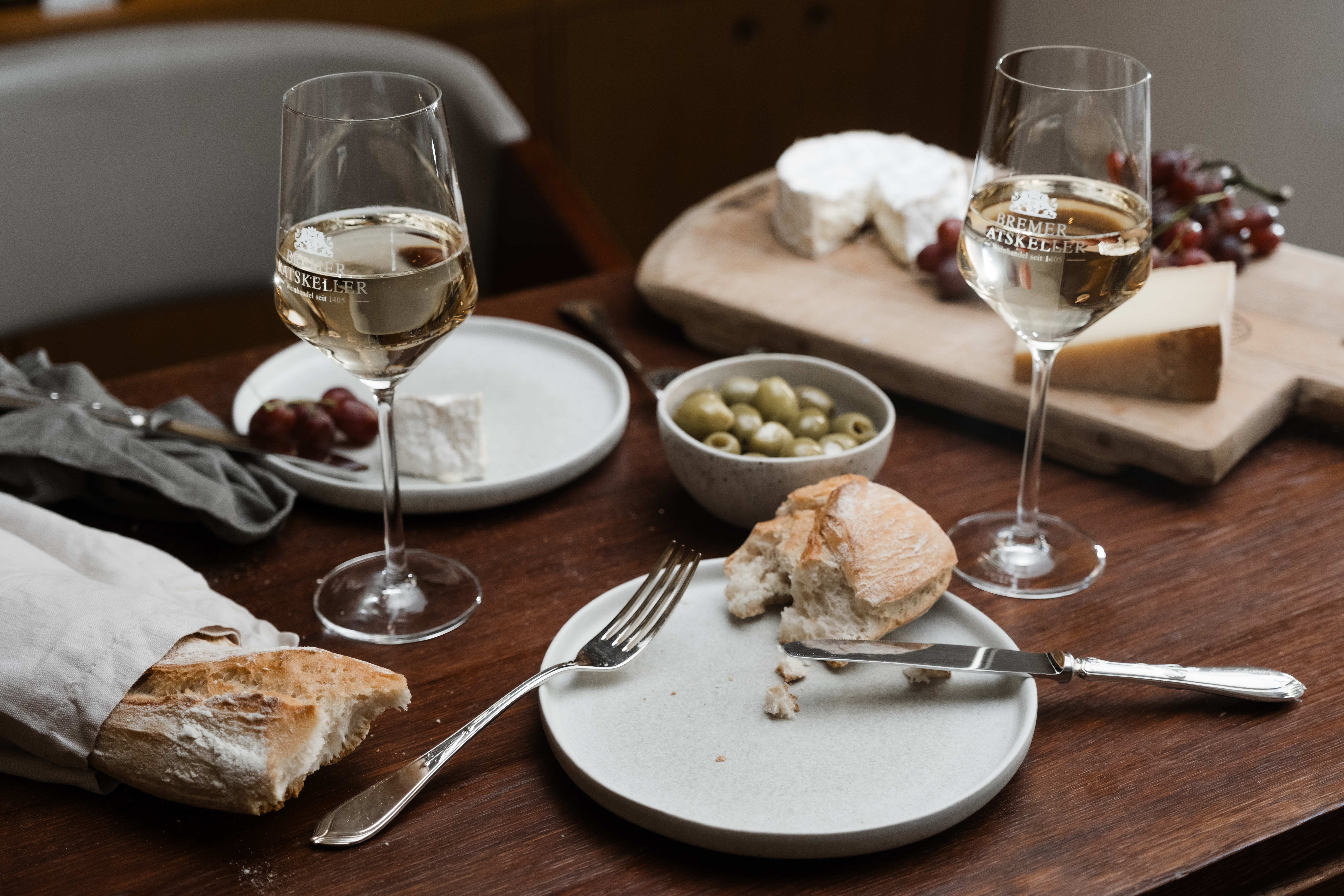 Holztisch mit zwei Weißweingläsern, Oliven, Käse und Baguette